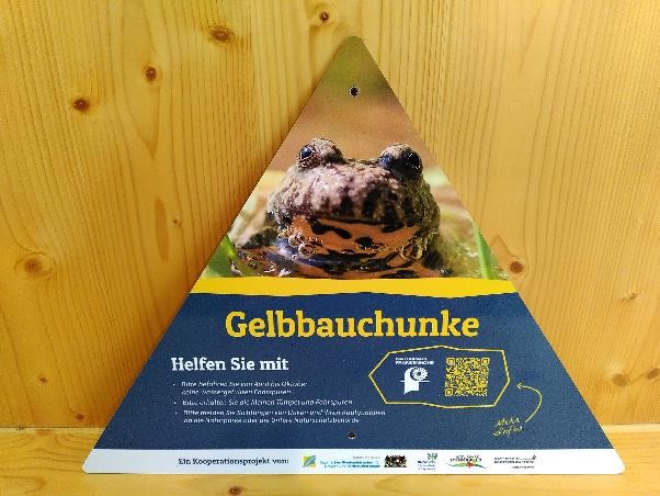 Ein Schild mit einer Gelbbauchunke, das daraufhinweißt, dass Wanderer und Fahrradfahrer die Tiere nicht gefährden sollen.